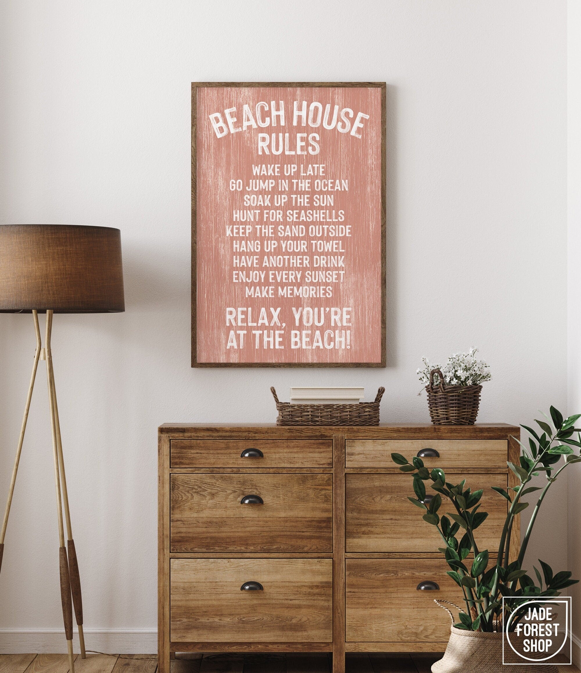 The Beach House Rules Sign, Painted Wood, Coral, adds charm as beach house wall decor when displayed above a wooden dresser next to a lamp and a basket of flowers.