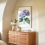 a wooden dresser with a framed picture of purple hydrangeas hanging on the wall above it. The dresser has six drawers and a vase of white flowers on top.
