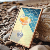 A framed illustration of a cocktail glass with orange slices is displayed on a rocky beach.