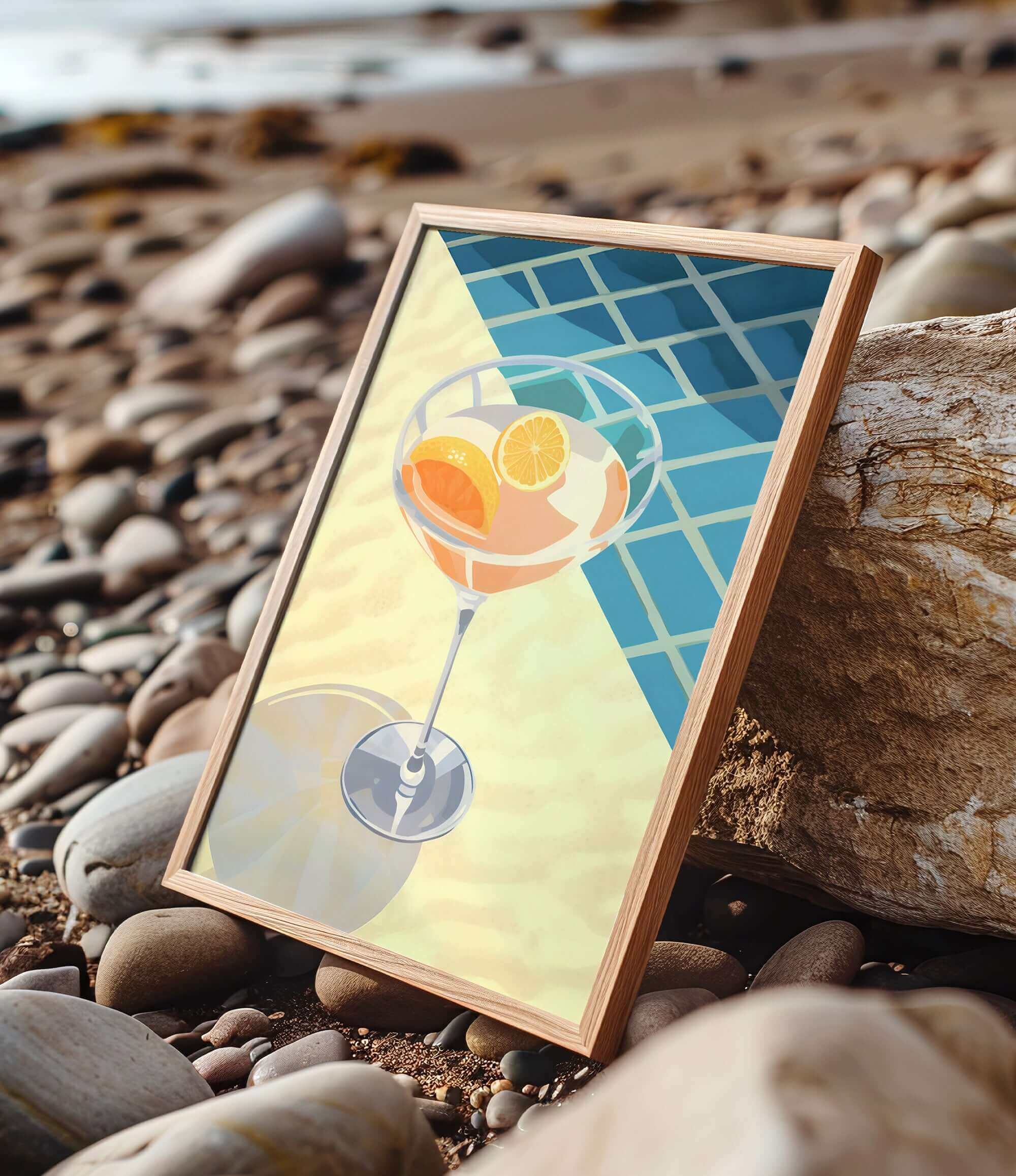 A framed illustration of a cocktail glass with orange slices is displayed on a rocky beach.