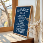 A framed sign on a window sill that reads "LAKESHOUSE RULES" and provides a list of activities to be enjoyed at the lake, including "WAKE UP LATE", "GO JUMP IN THE LAK", "SINK IN THE LAK", "TAKE A NAP", "SIP A DRINK", "FEEL THE BREZEF", "ENJOY EVERY SUNSET", and "MAKE MEMORIES".