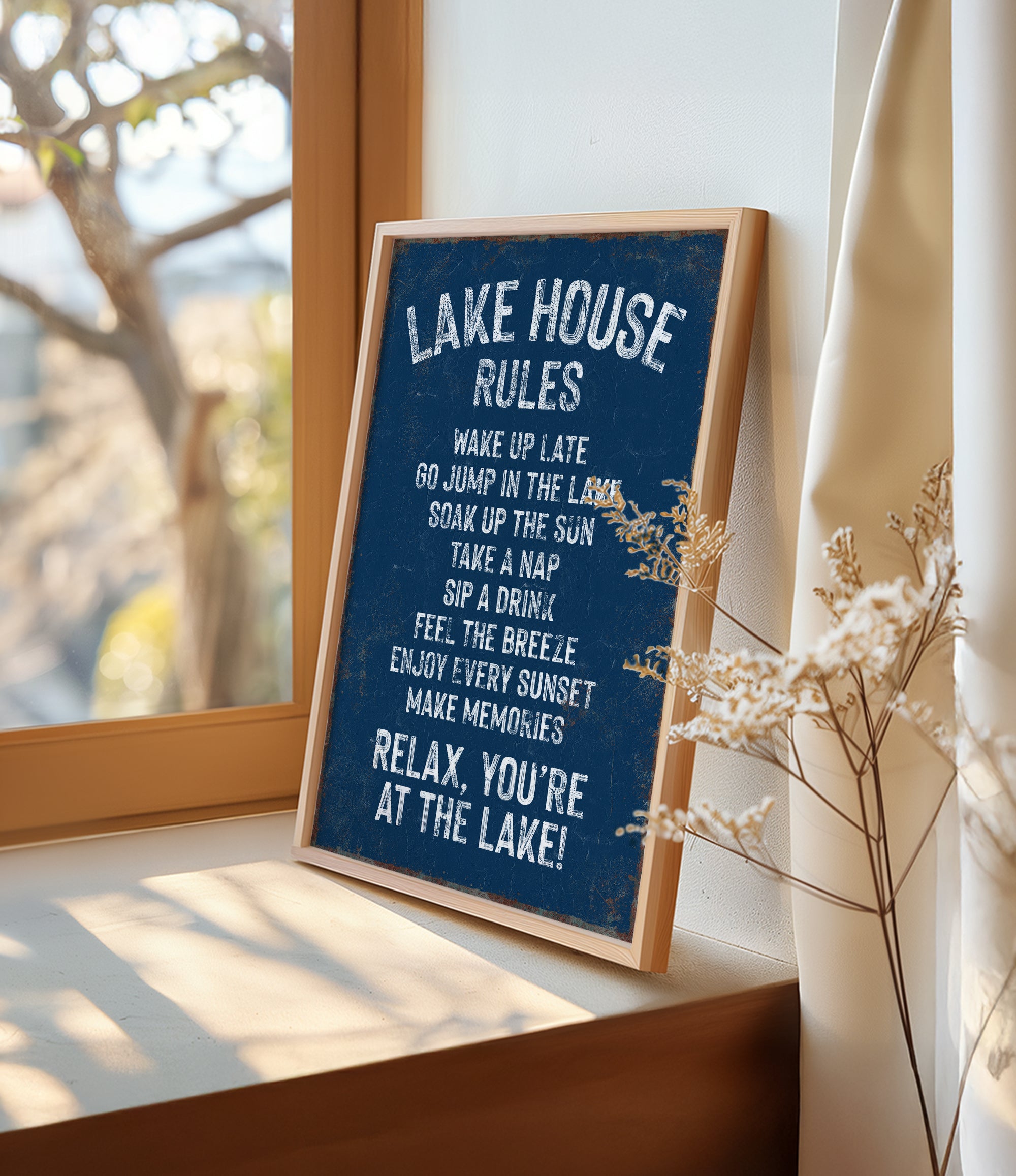 A framed sign on a window sill that reads "LAKESHOUSE RULES" and provides a list of activities to be enjoyed at the lake, including "WAKE UP LATE", "GO JUMP IN THE LAK", "SINK IN THE LAK", "TAKE A NAP", "SIP A DRINK", "FEEL THE BREZEF", "ENJOY EVERY SUNSET", and "MAKE MEMORIES".