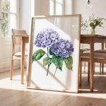 a framed painting of purple hydrangeas on a wooden table, with a vase of greenery and a wooden chair visible in the background.