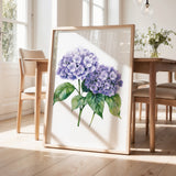 a framed painting of purple hydrangeas on a wooden table, with a vase of greenery and a wooden chair visible in the background.