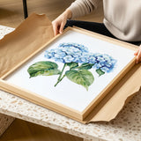 A person is holding a framed painting of blue hydrangea flowers in a wooden frame.