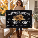 A woman is holding a large sign that reads "Autumn Harvest Flower Shop" with a floral design.