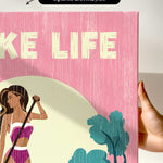 a hand holding a pink canvas print with a stylized illustration of a woman in a bikini.