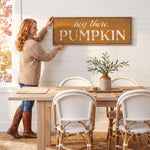 A woman hangs a framed sign on a wall above a wooden dining table with white chairs.