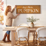 A woman hangs a framed sign on a wall above a wooden dining table with white chairs.