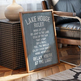 A framed sign on a wooden floor next to a chair, with a lamp and a black and white rug in the background.