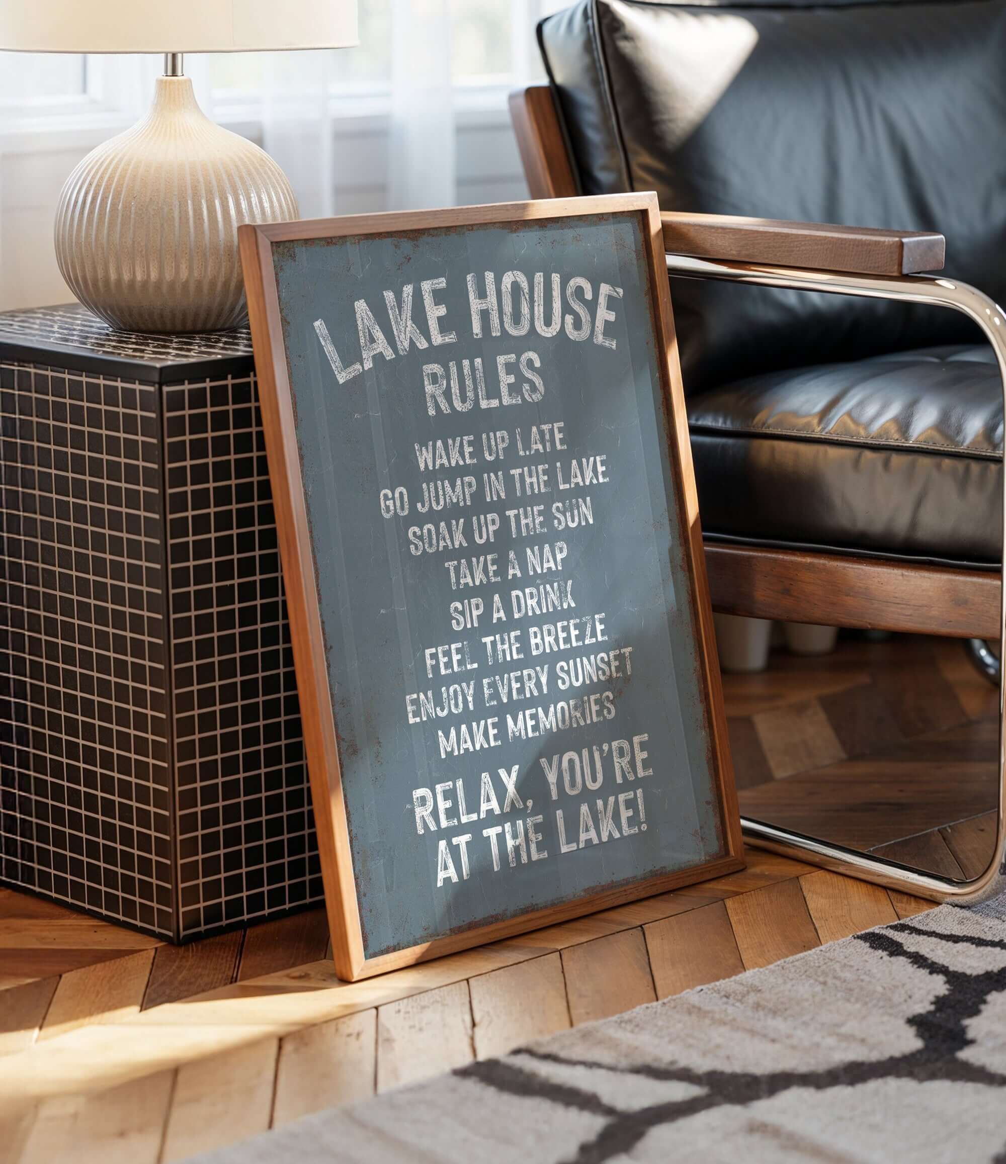 A framed sign on a wooden floor next to a chair, with a lamp and a black and white rug in the background.