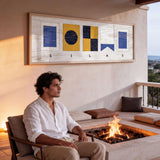A man sits on a chair in front of a fireplace, with a framed artwork hanging on the wall behind him.