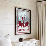 a framed poster of a mountain landscape with two red Adirondack chairs and skis, hanging on a wall above a white dresser.