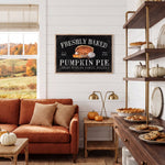 a cozy living room with a large window overlooking a picturesque autumn landscape. The room is decorated with a rustic wooden coffee table, a comfortable sofa, and a wooden side table with a lamp. A framed sign on the wall announces "Freshly Baked Pumpkin Pie", and the room is filled with various baked goods, including pies and cookies, suggesting a festive atmosphere.