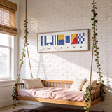 a cozy bedroom with a wooden bed suspended from the ceiling by ropes, creating a unique and comfortable sleeping space. The bed is adorned with white pillows and a pink blanket, adding a touch of warmth and comfort to the room. Above the bed, there is a framed artwork depicting nautical flags, adding a nautical theme to the space. The room is also decorated with potted plants, which contribute to the overall pleasant and inviting atmosphere.