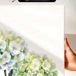 a hand holding a white canvas print with a watercolor painting of white hydrangea flowers on it.
