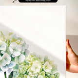 a hand holding a white canvas print with a watercolor painting of white hydrangea flowers on it.