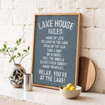 A framed sign with a list of rules for a lake house, accompanied by a potted plant, a metal container, and a bowl of lemons, all set against a white brick wall.