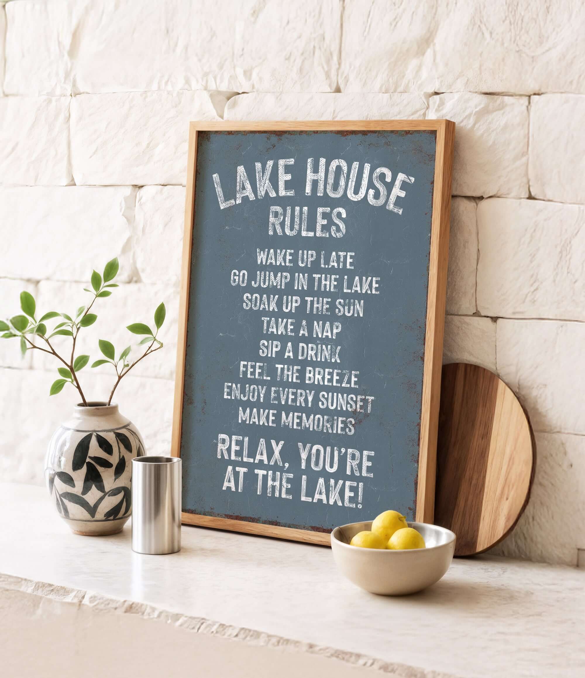A framed sign with a list of rules for a lake house, accompanied by a potted plant, a metal container, and a bowl of lemons, all set against a white brick wall.