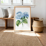 a framed painting of blue hydrangea flowers on a wooden bench, with a woven basket and a woven rug on the floor in the background.