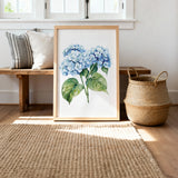 a framed painting of blue hydrangea flowers on a wooden bench, with a woven basket and a woven rug on the floor in the background.