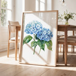 a framed painting of blue hydrangea flowers on a wooden table, with a vase of greenery and a wooden chair visible in the background.