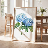 a framed painting of blue hydrangea flowers on a wooden table, with a vase of greenery and a wooden chair visible in the background.