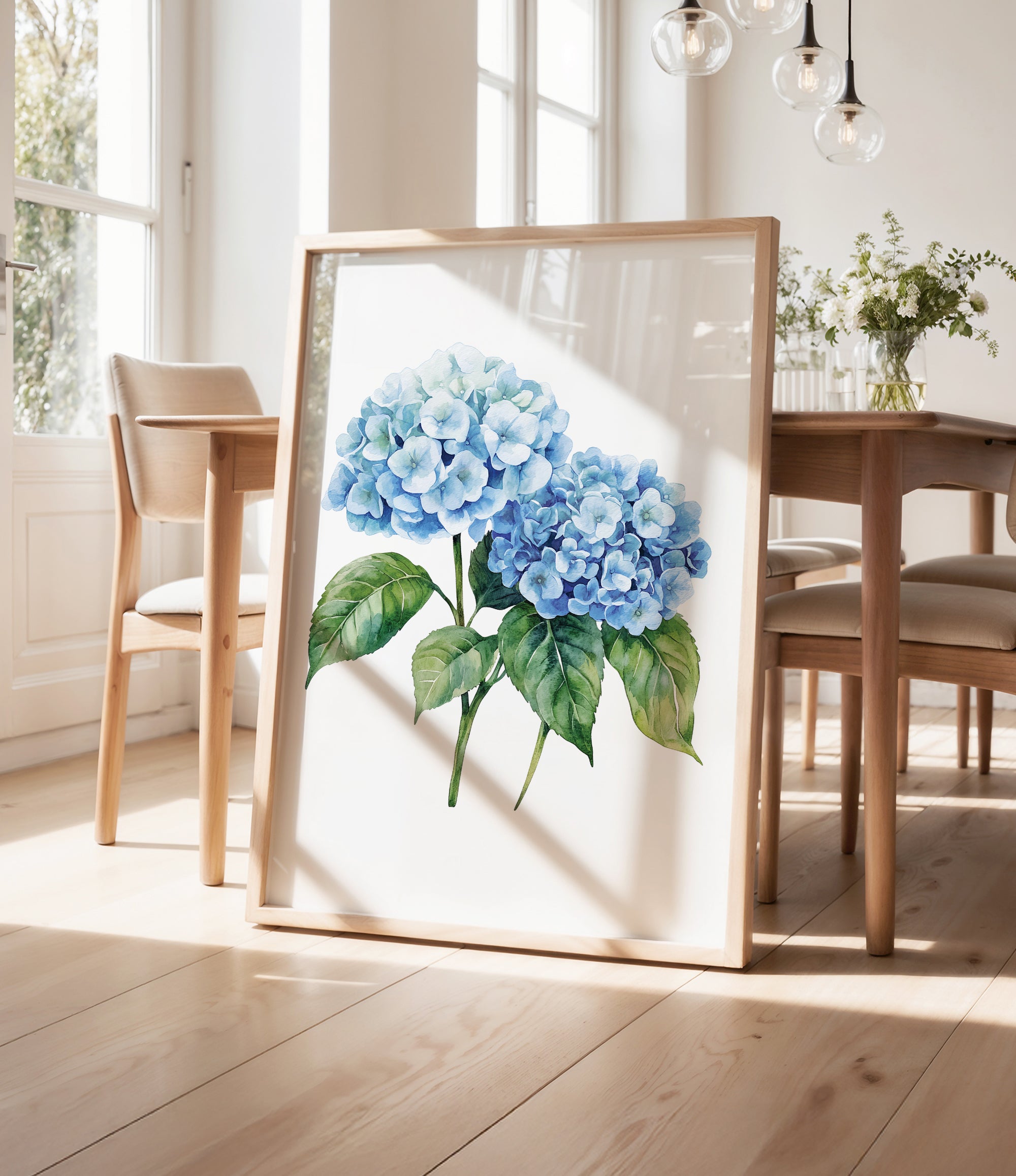 a framed painting of blue hydrangea flowers on a wooden table, with a vase of greenery and a wooden chair visible in the background.