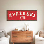 a living room with a large red sign that says "APRÈS SKI" above a wooden dresser. The dresser has a vase of flowers and a book on it. There are two white chairs in the room, and a small table with a lamp on it.