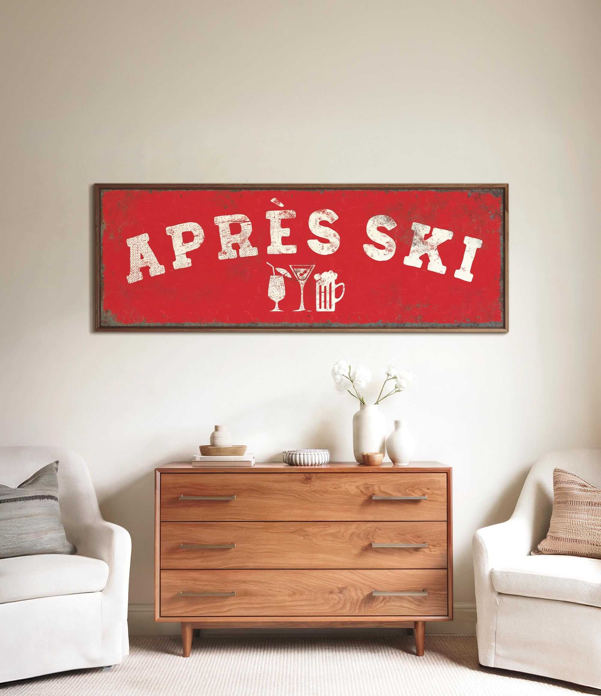a living room with a large red sign that says "APRÈS SKI" above a wooden dresser. The dresser has a vase of flowers and a book on it. There are two white chairs in the room, and a small table with a lamp on it.