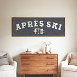 a wooden dresser with a framed sign above it that says "APRÈS SKI", and there are two white chairs and a vase with flowers placed in front of the dresser.