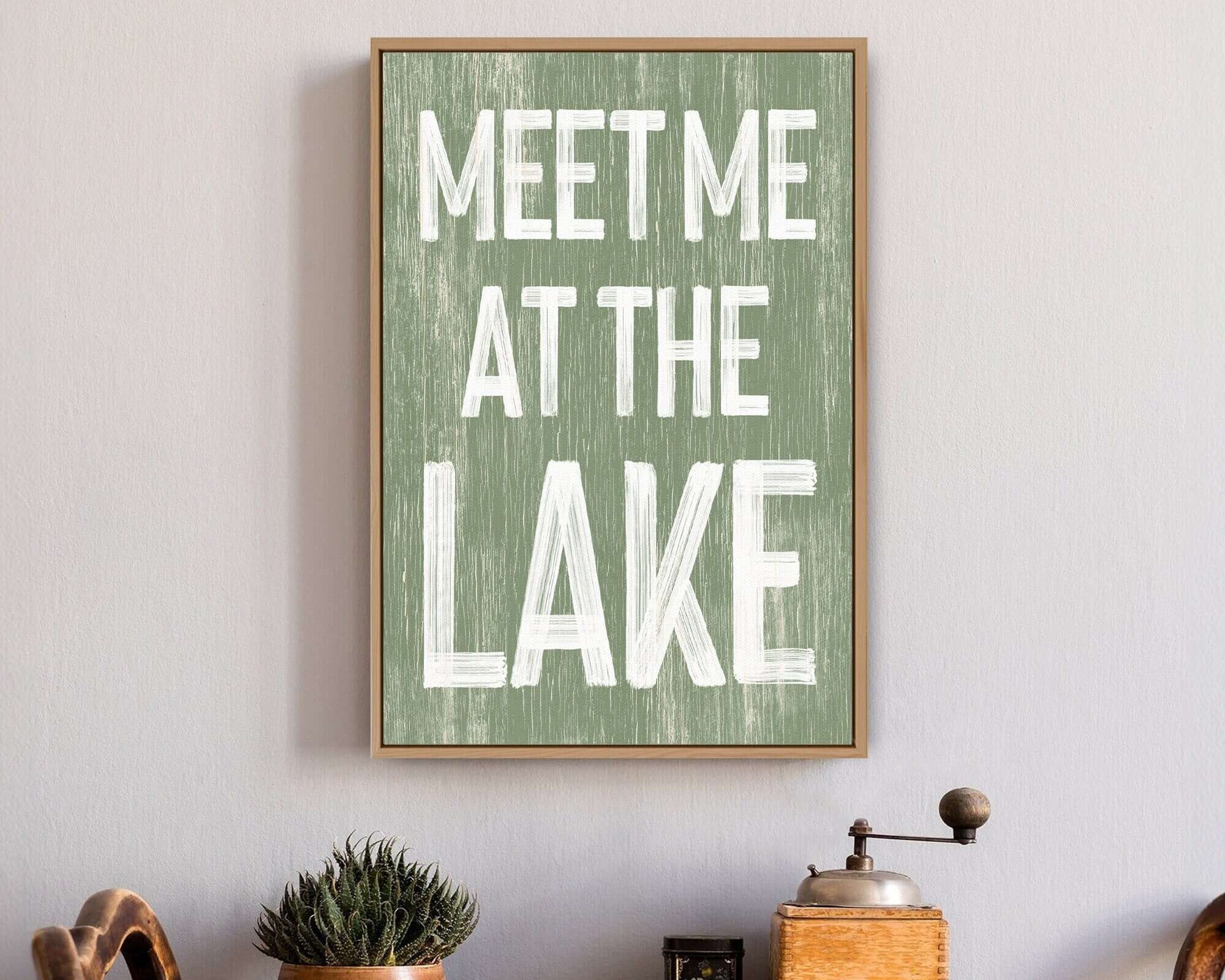 The "Meet Me at the Lake Sign, Seagrass" is a framed retro sign that reads "MEET ME AT THE LAKE," ideal above a table with plants and kitchen items for charming lake house decor.