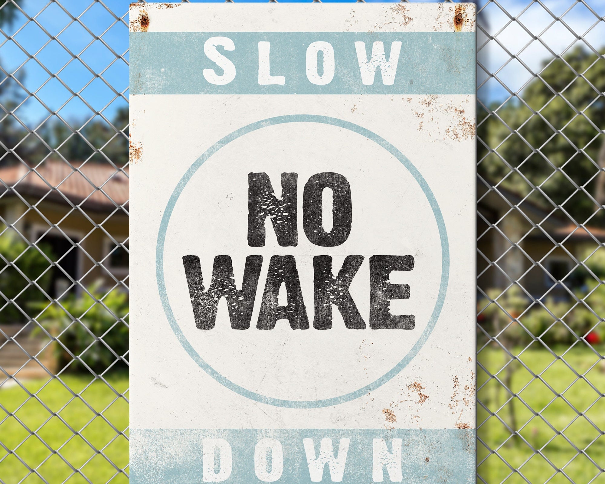 The No Wake Sign, Tide on White, features a vintage design—reading "SLOW NO WAKE DOWN"—against a chain-link fence with grass and a house in the background, perfect for lake house wall decor.