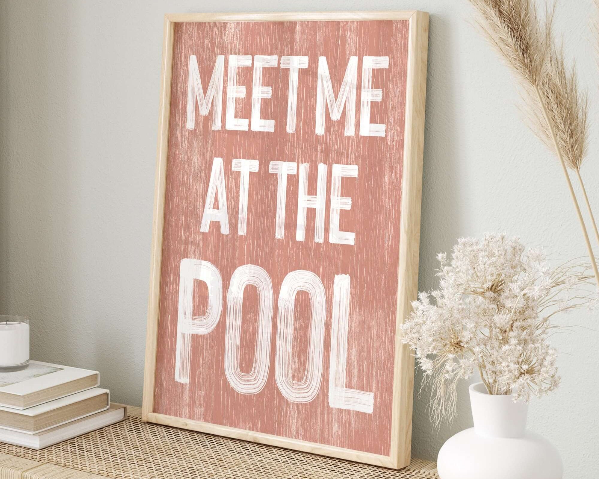 The Meet Me at the Pool Sign, Coral, is framed and displayed on a woven table with books and dried flowers, bringing a modern farmhouse vibe to any space.