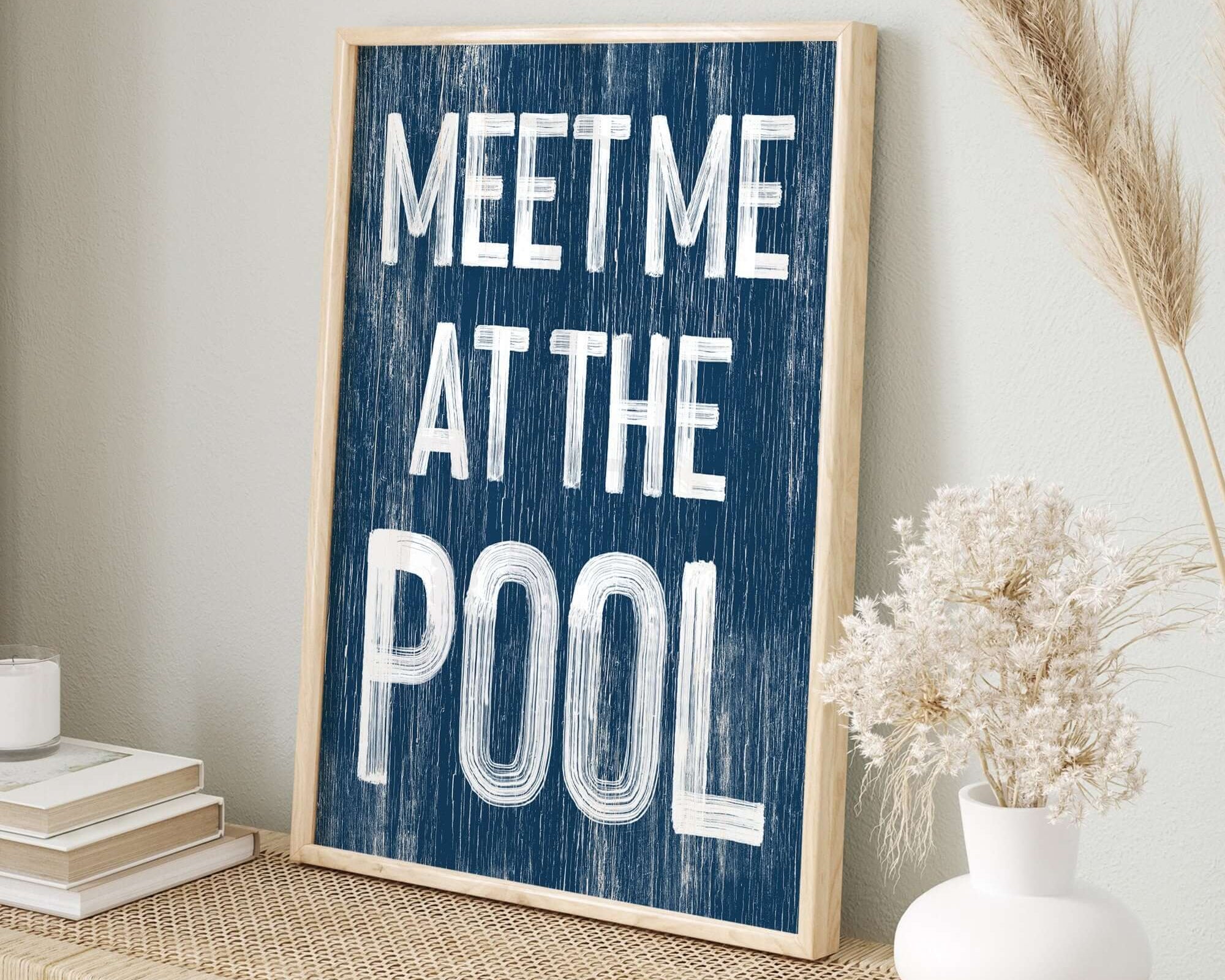 The Meet Me at the Pool Sign, Nautical, is framed and displayed on a woven shelf with books and a vase of dried grass, bringing retro charm to your modern farmhouse décor.