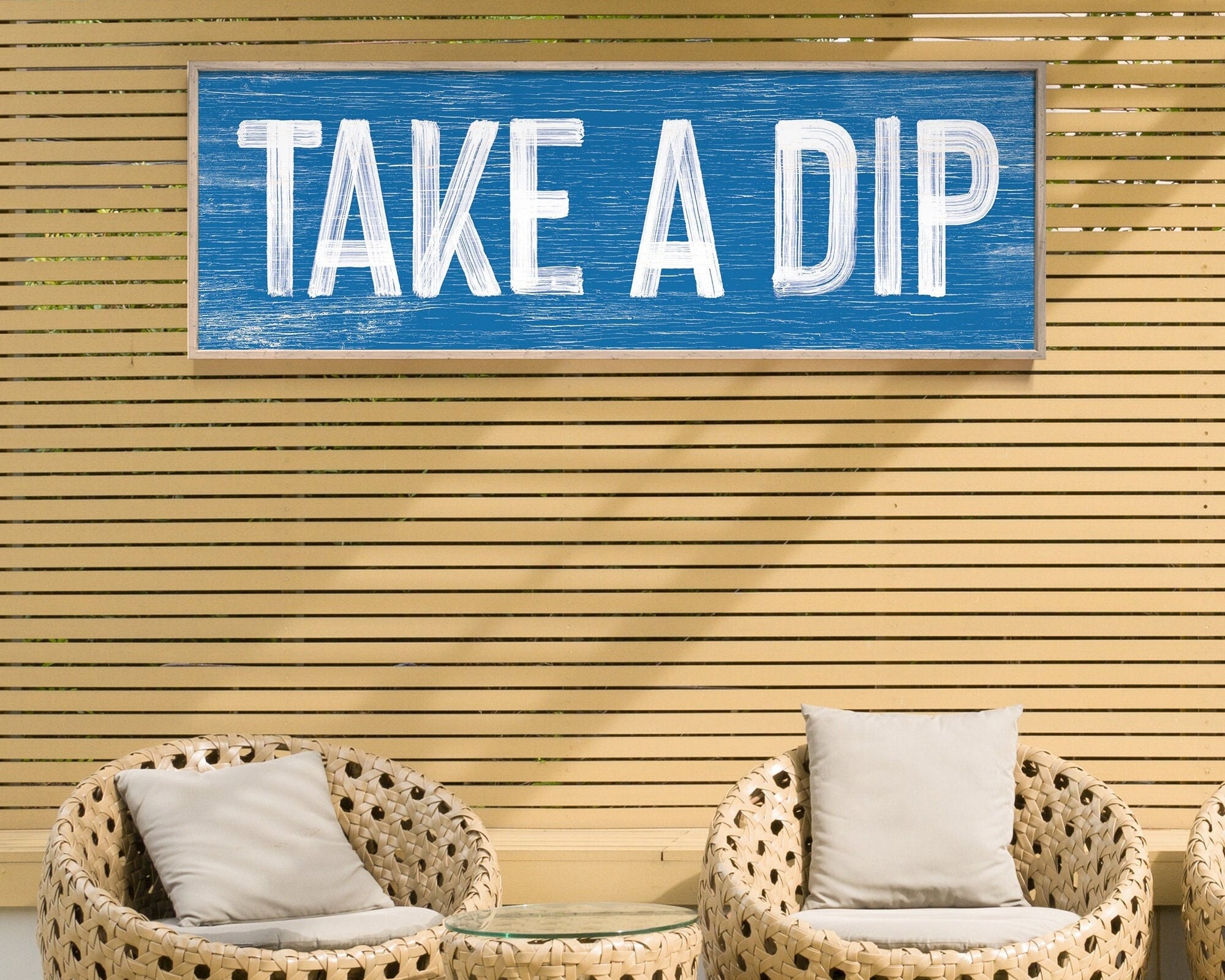 Two wicker chairs and a table sit under a Take a Dip Sign in Ocean Blue against wooden slats, bringing modern farmhouse charm to this inviting outdoor space.