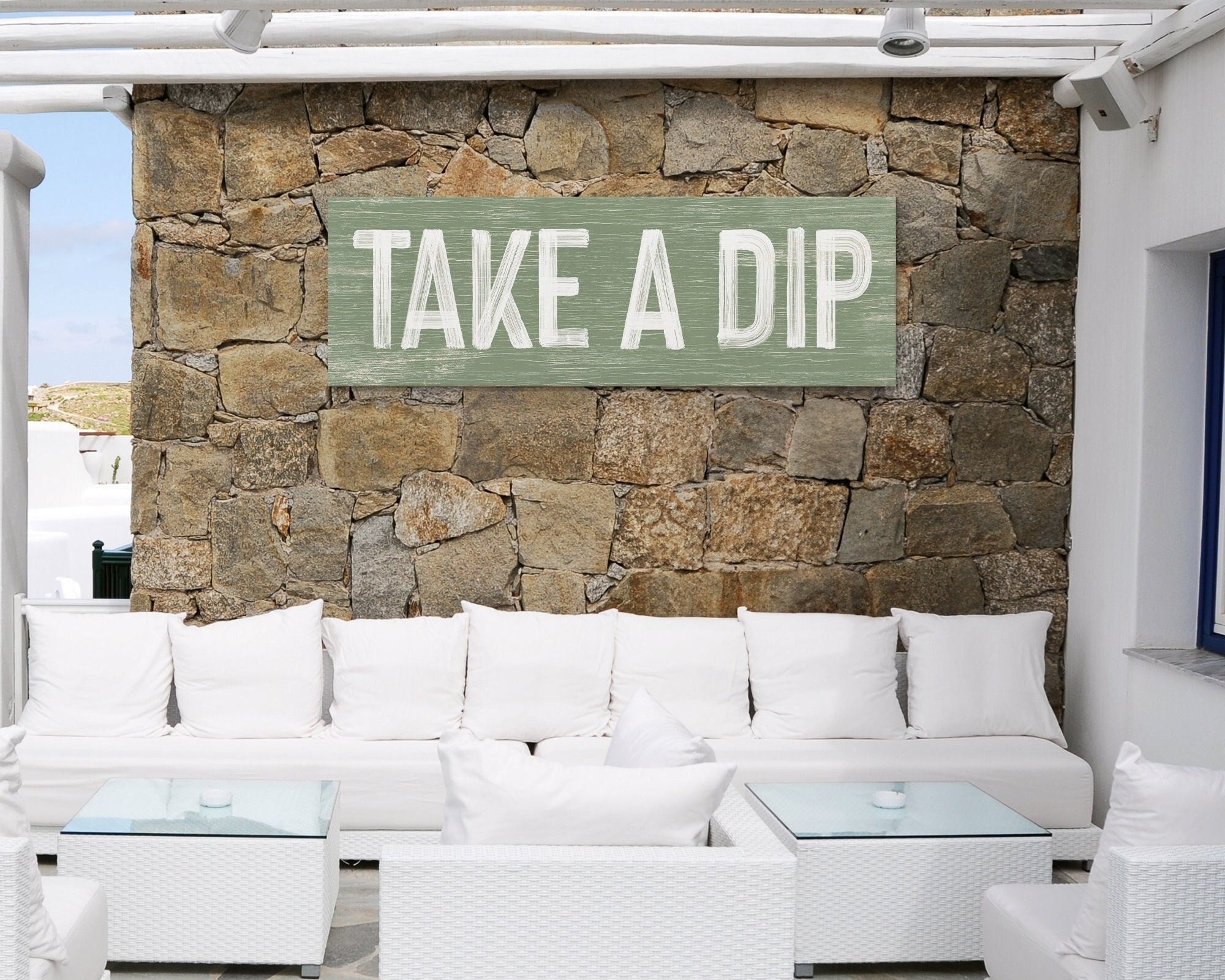 Outdoor living space featuring white sofas, glass tables, and a stone wall adorned with the stylish Take a Dip Sign in Seagrass—perfect for modern farmhouse decor lovers.