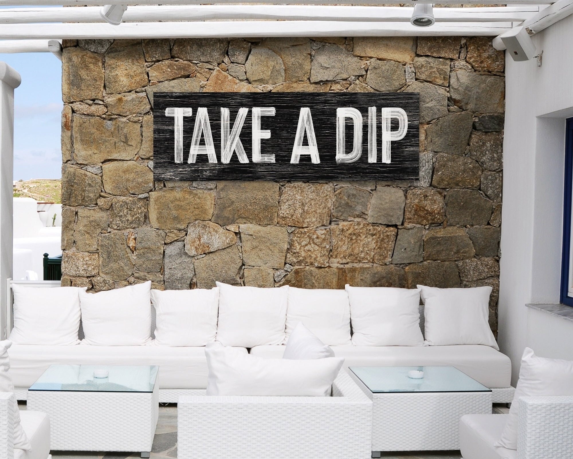 Outdoor lounge area with white sofas, glass tables, and a stone wall showcases modern farmhouse decor, highlighted by the "Take a Dip Sign, Black" above the seating.