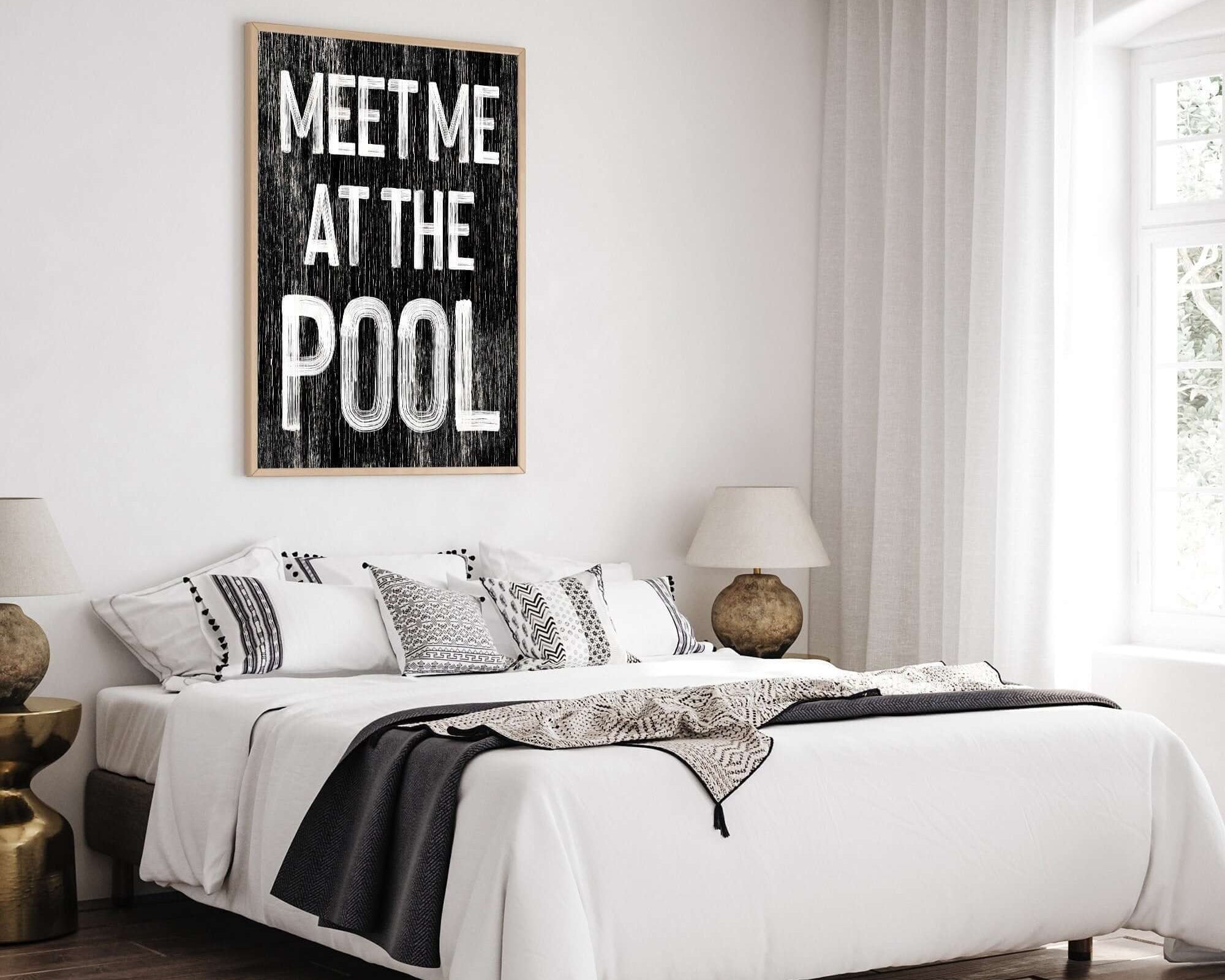 A cozy bedroom featuring a bed, decorative pillows, and the "Meet Me at the Pool Sign, Black" as vintage wall decor enhances the summer home aesthetic.