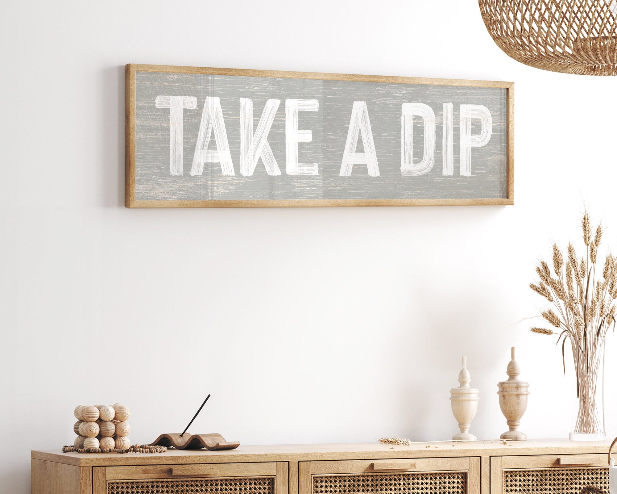 A Take a Dip Sign in stone hangs above a rattan cabinet in a bright, airy room with natural accents, adding farmhouse charm to your indoor or outdoor space.