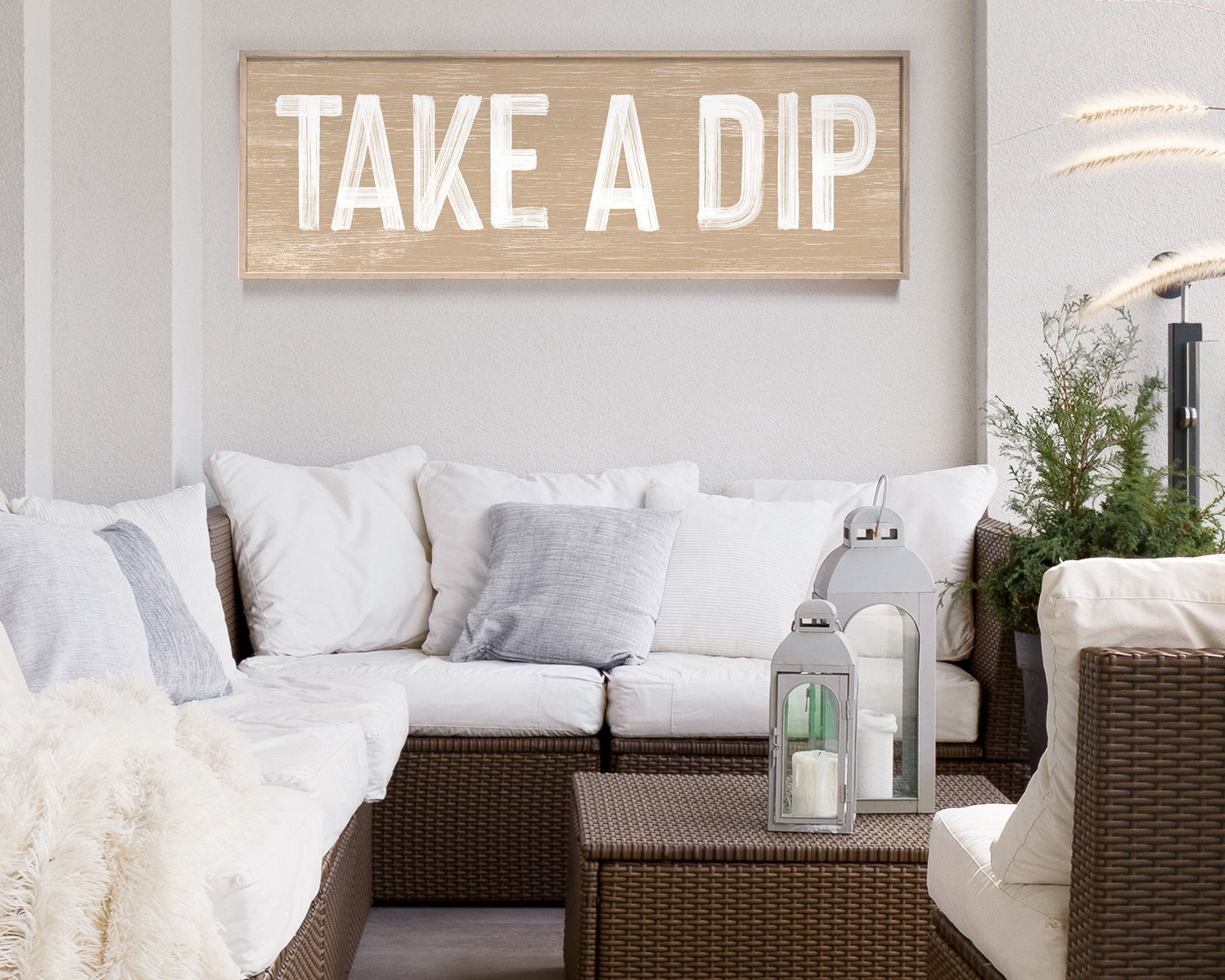 Outdoor wicker sofa with cushions and a Take a Dip Sign, Latte on the wall; lanterns and plants add charm to this inviting modern farmhouse outdoor living space.