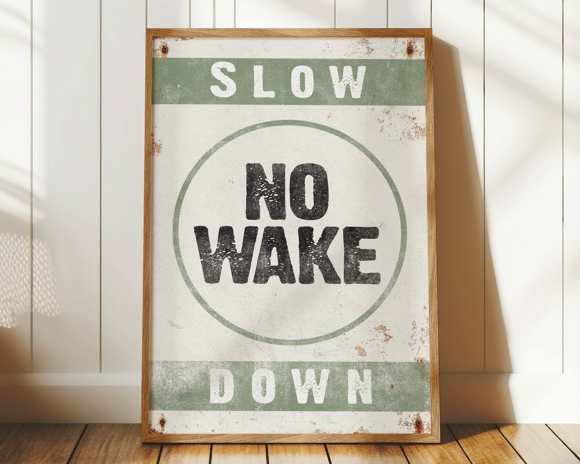 The No Wake Sign, Seagrass on White, is a framed vintage piece featuring "Slow Down No Wake" and rests on a wood floor against a white paneled wall—ideal lake house decor for your modern farmhouse style.