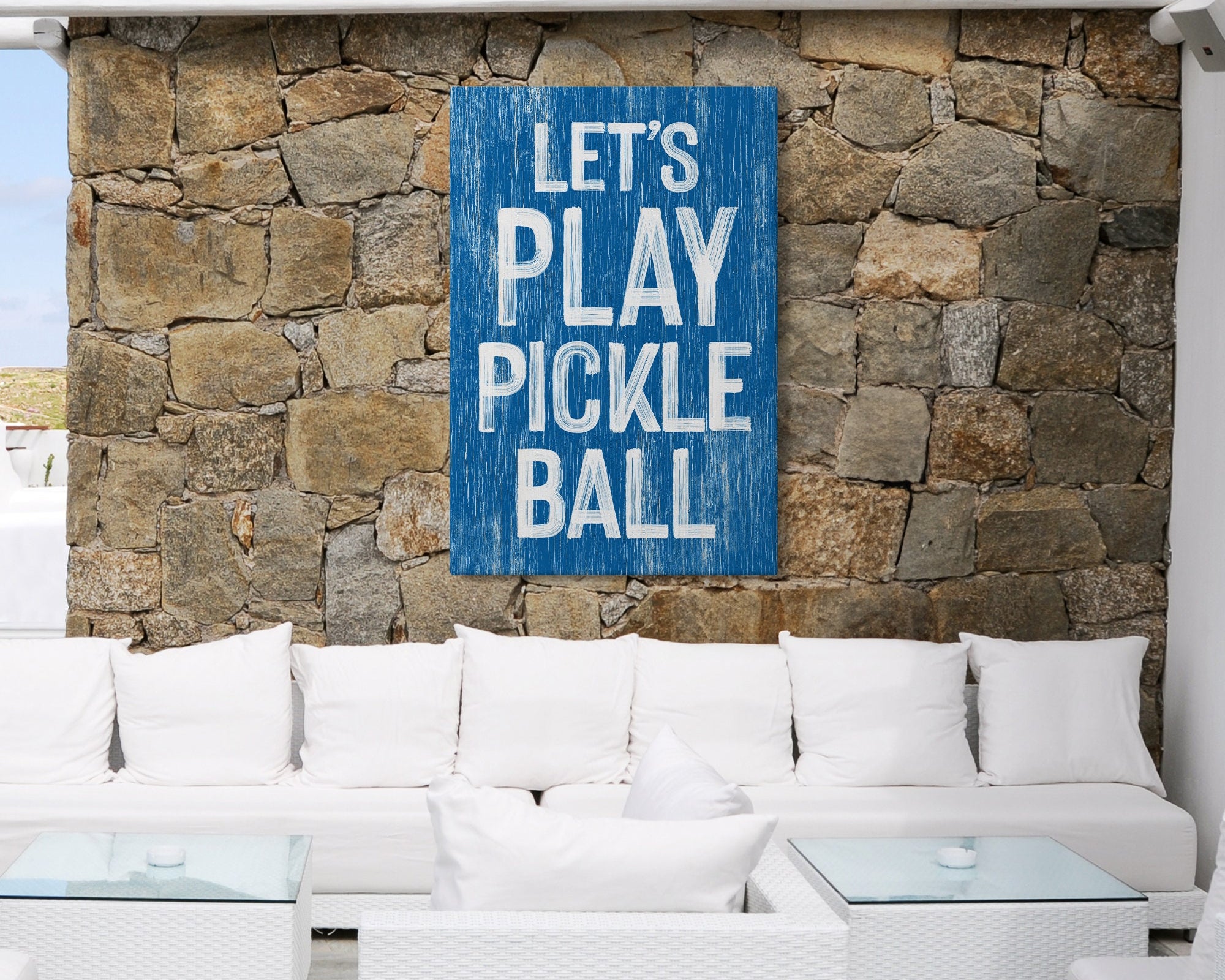 White outdoor lounge with a stone wall featuring the "Let's Play Pickleball Sign, Ocean Blue" sports-themed wall art.