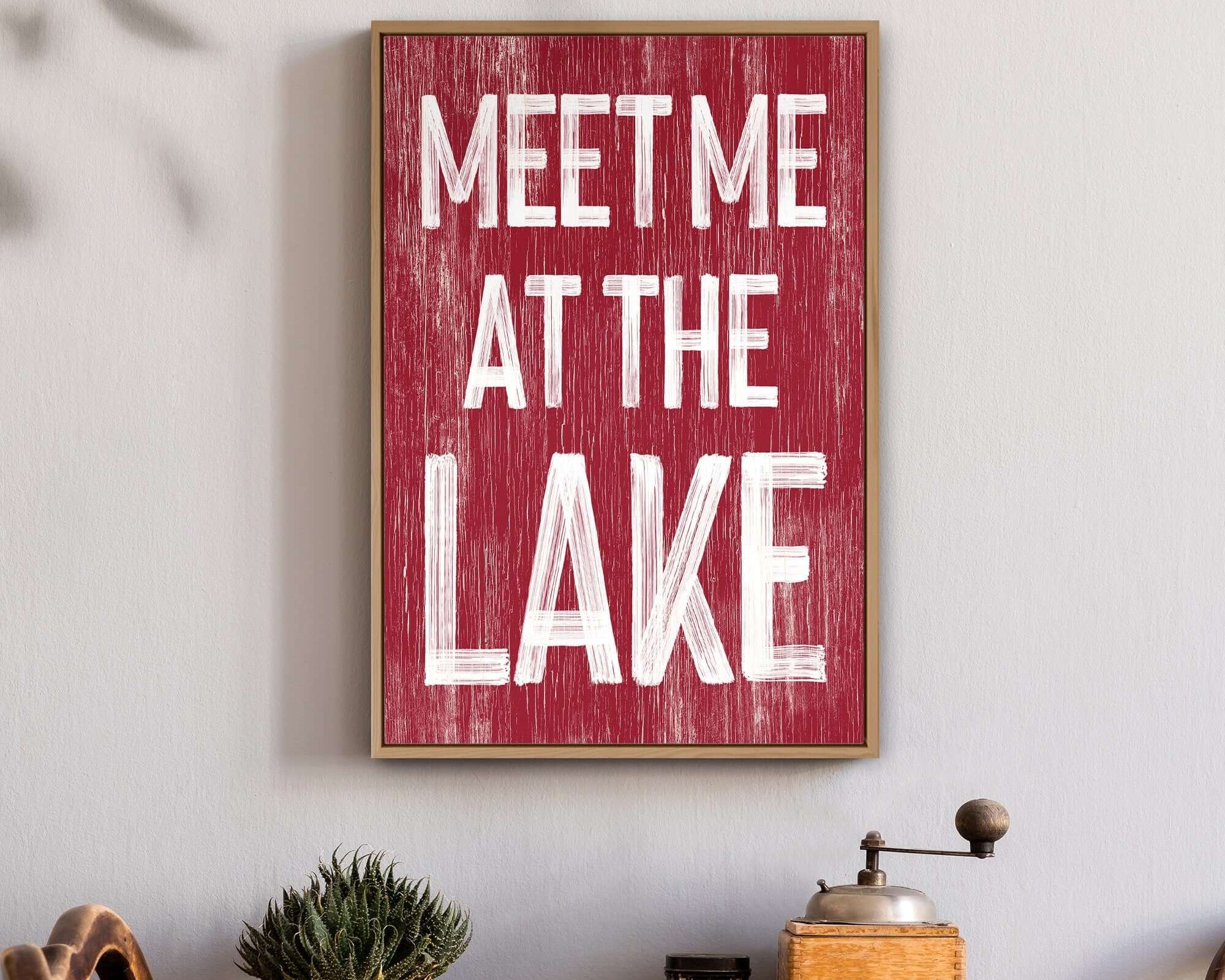 A Meet Me at the Lake Sign in Dark Red hangs above a table with plants and kitchen items, bringing charming lake house decor to the space.