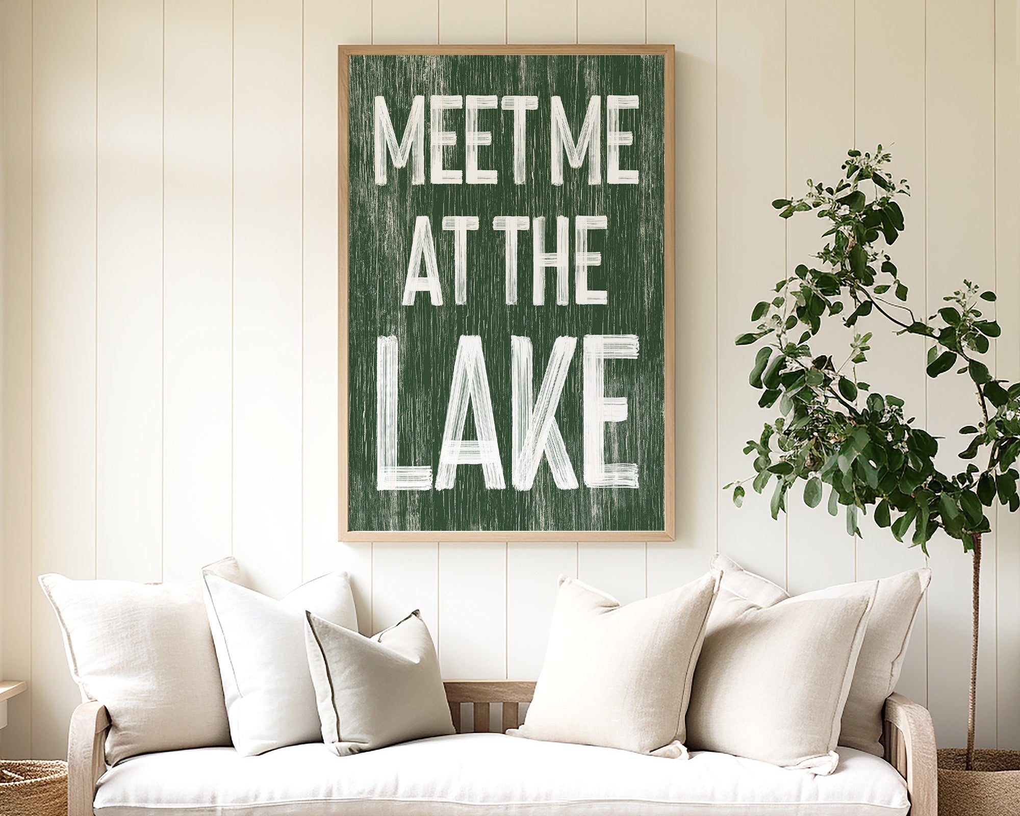 The "Meet Me at the Lake Sign, Vine" hangs above a beige couch with white pillows and a potted plant, adding cozy lake house decor and modern farmhouse charm to the space.