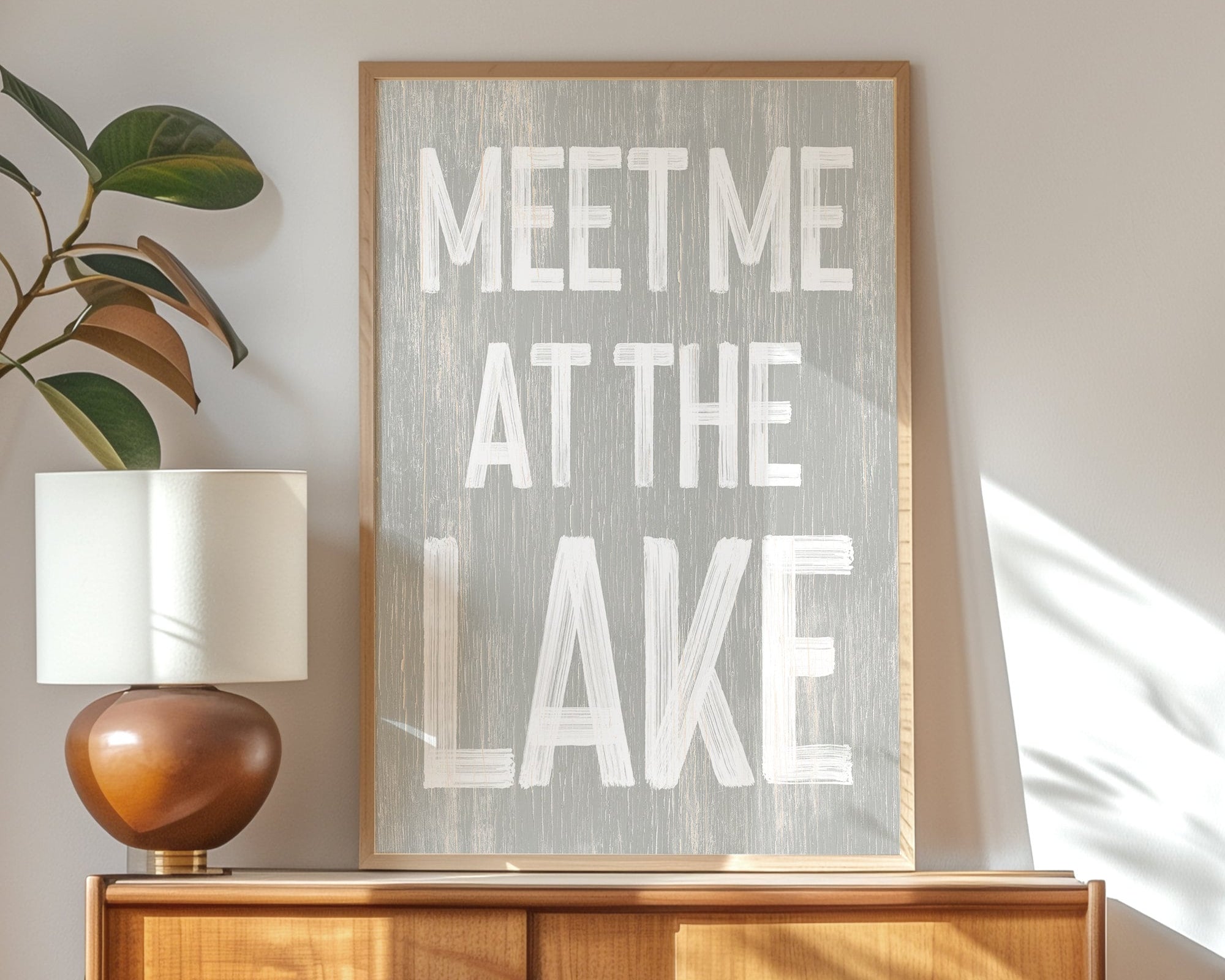 The Meet Me at the Lake Sign, Stone, is displayed on a wooden cabinet next to a lamp and plant—an ideal accent for your lake house decor.