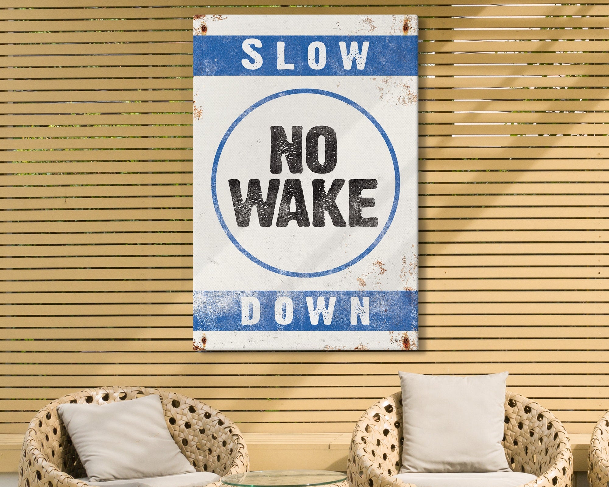 A vintage "No Wake Sign, Ocean Blue on White" hangs on a wooden slat wall above two wicker chairs with cushions, adding charming lake house decor.