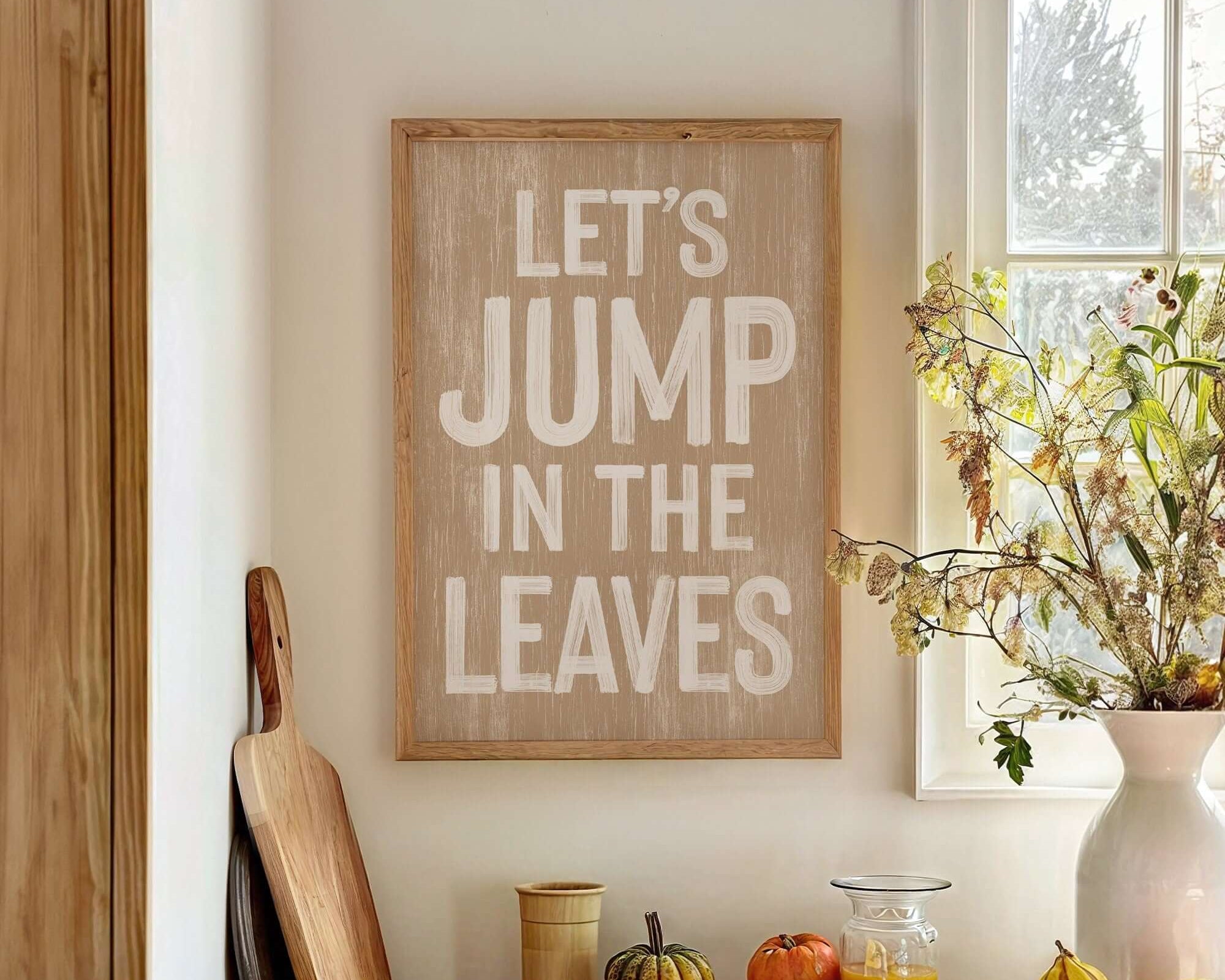 The Let's Jump in the Leaves Sign, Latte brings charm to your autumn farmhouse decor, displayed above fall wall accents and seasonal fruit on a bright kitchen counter.
