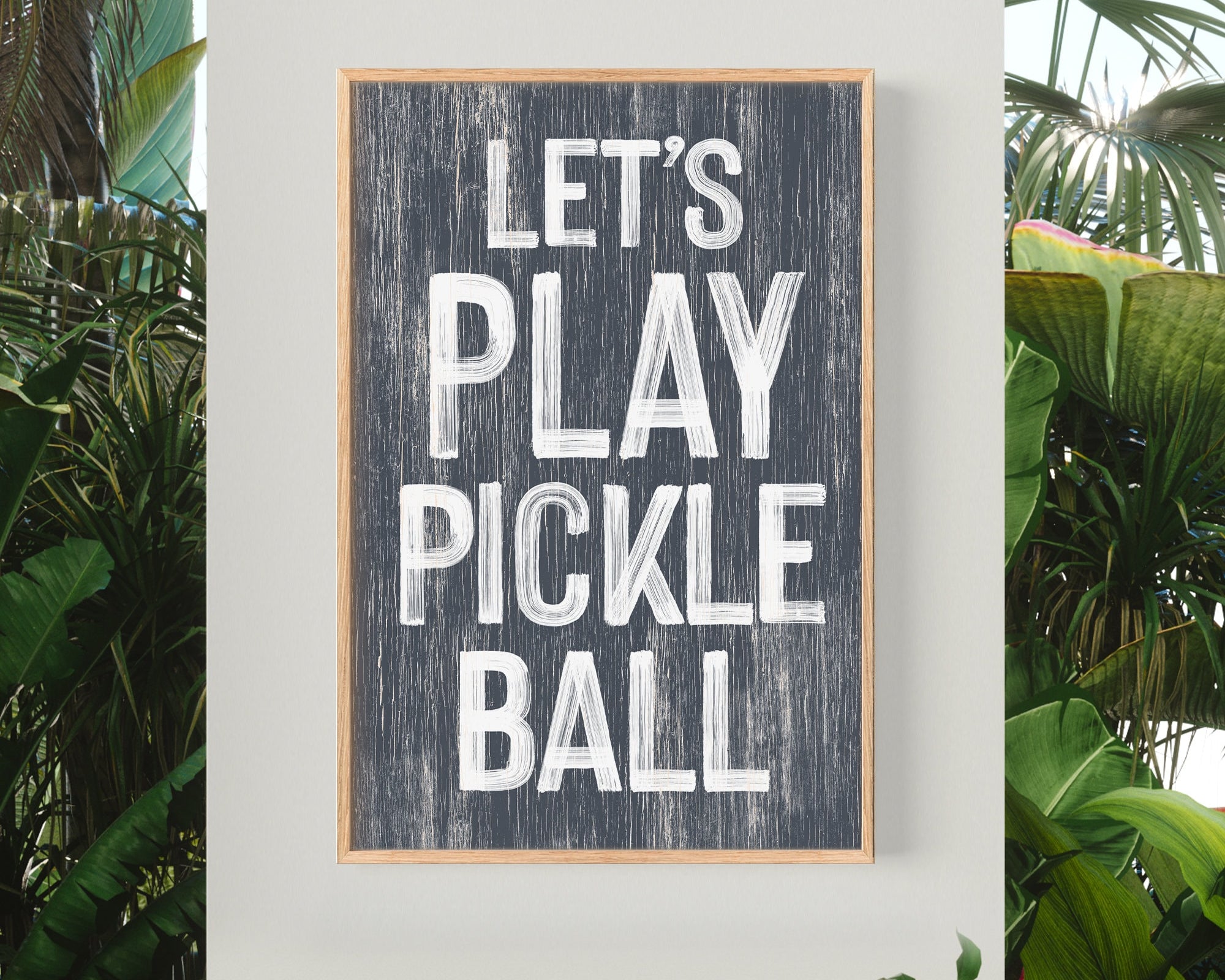 The Let's Play Pickleball Sign in Hale Navy hangs on a wall with tropical plants, making the perfect sports-themed pickleball wall decor.