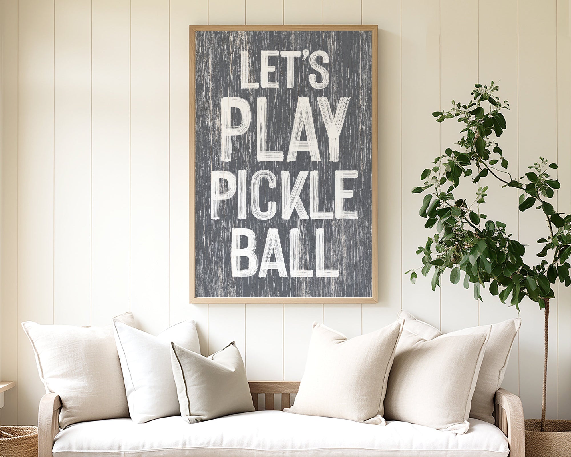 A cozy sofa sits beneath the "Let's Play Pickleball Sign, Slate" in a bright, airy room with modern farmhouse decor and a lively plant.